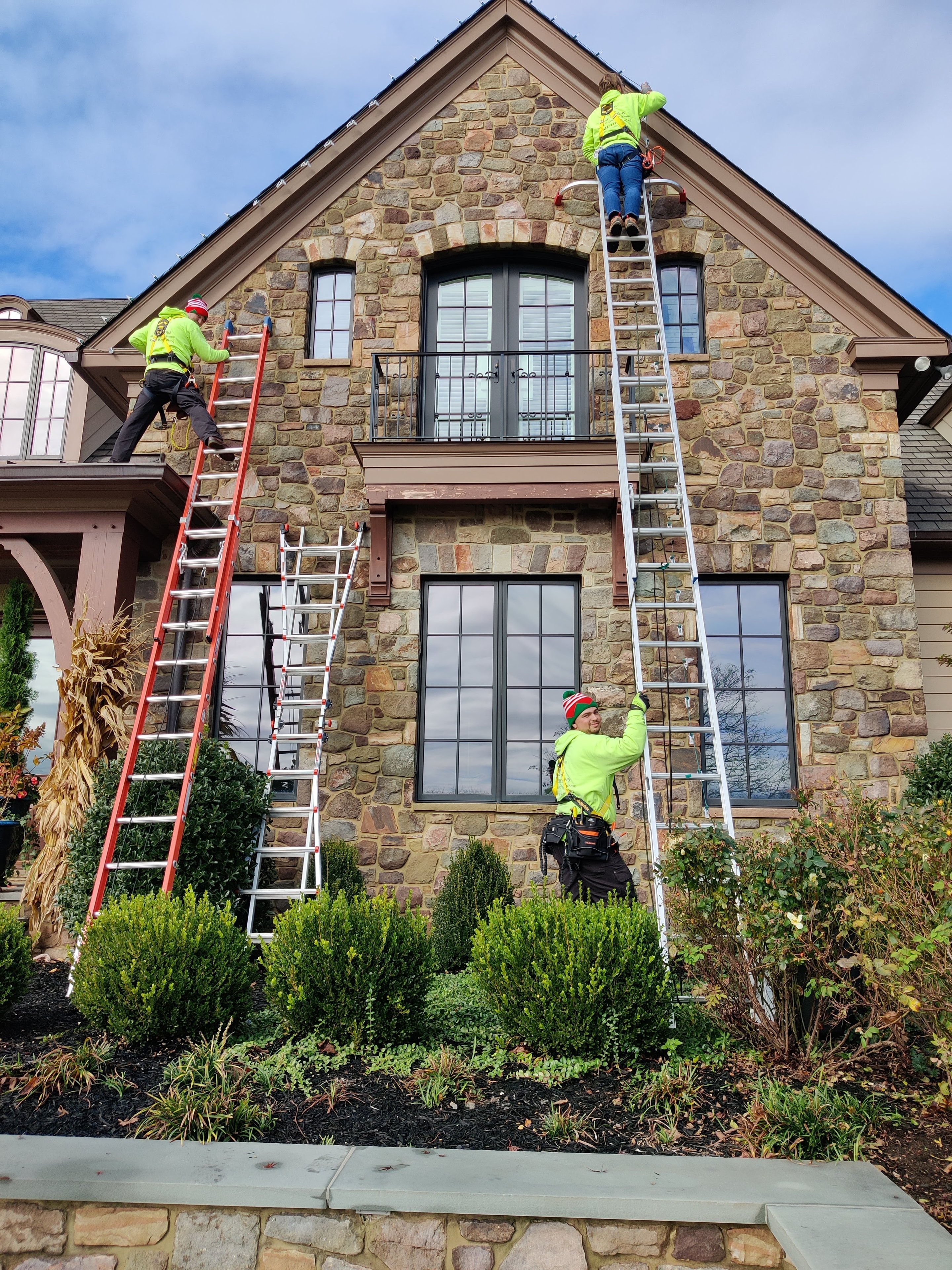 E.L.F. Designs crew with multiple ladders installing holiday lights on a large stone home in Churchville, Maryland
