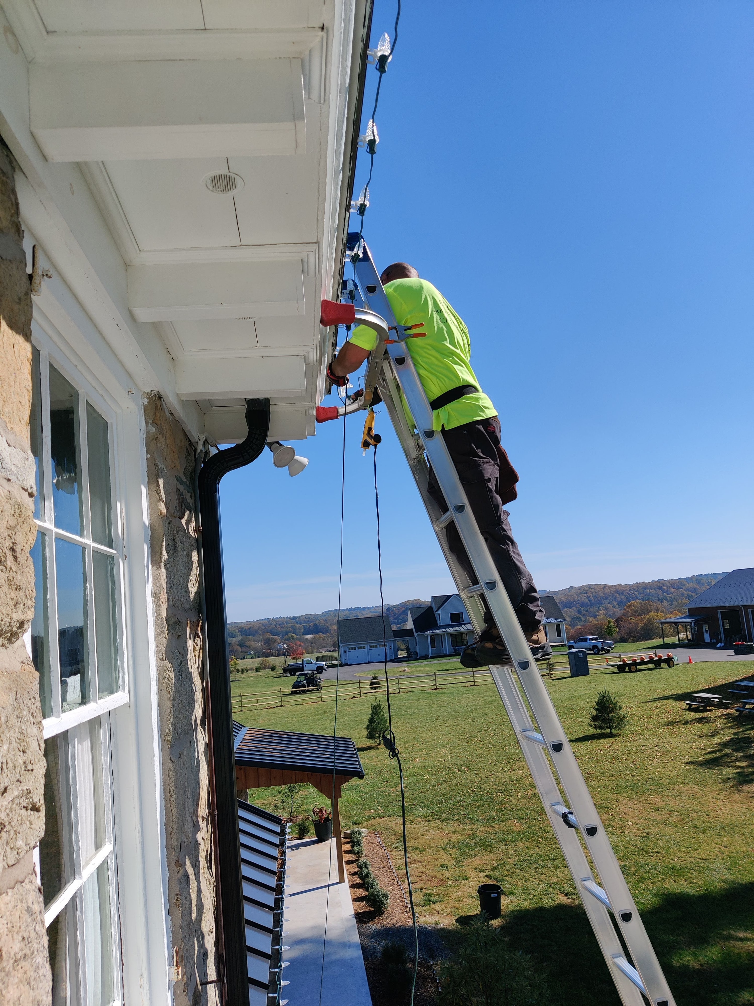 E.L.F. Designs technician on a ladder installing Christmas lights on a stone farmhouse in Jarrettsville, Maryland